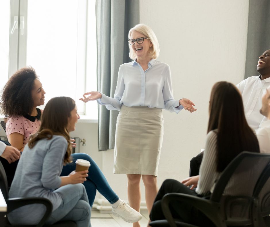 A cheerful woman presenting to a group in a casual team meeting setting.