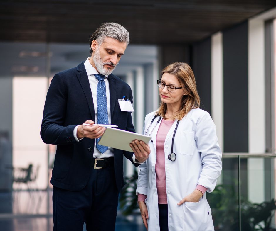 A businessman and a female doctor reviewing notes on a clipboard in a hospital corridor.