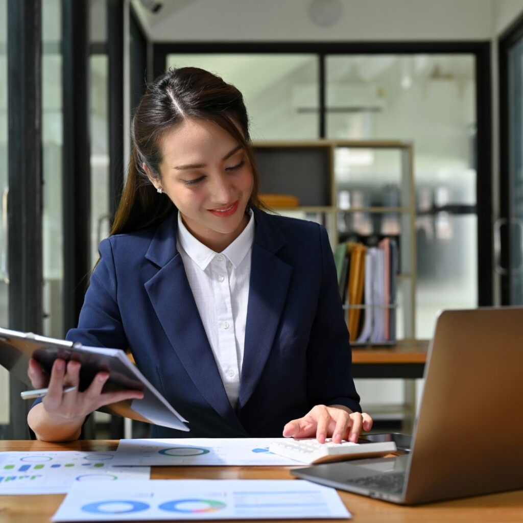 Businesswoman analyzing financial charts and working on a laptop at a desk.