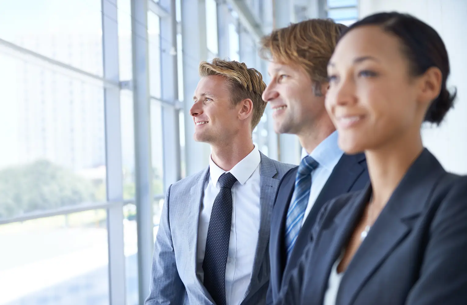 Three business professionals smiling while looking out of a modern office window.